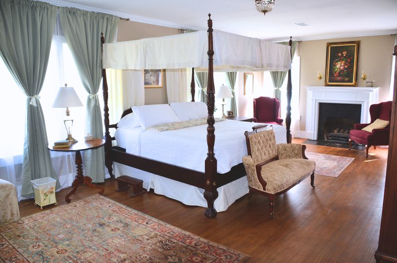The Thompson suite: a dark wood four-poster canopy bed with white linens, a tufted slipper chair, and a white fireplace flanked by burgundy wingback chairs.