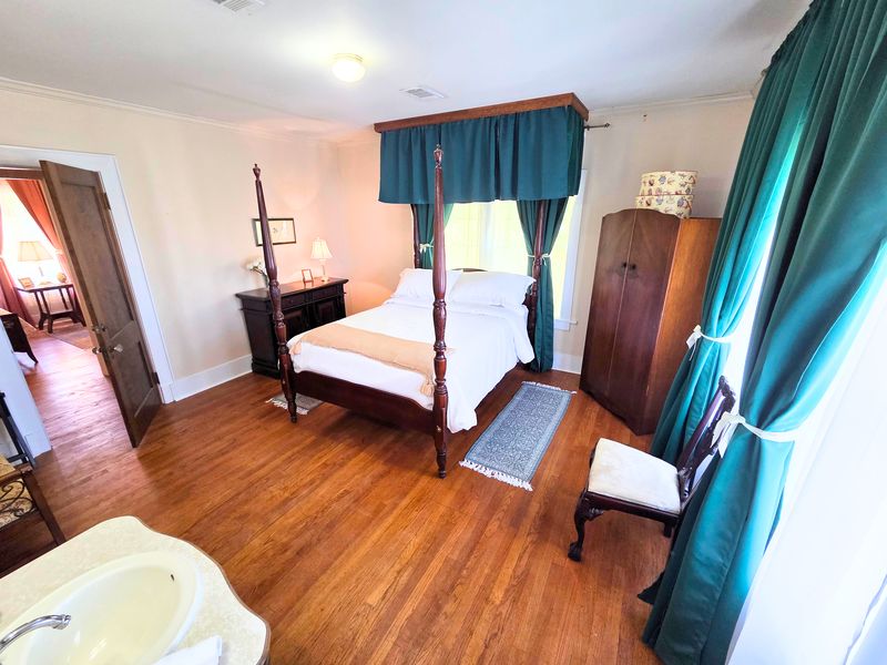 The Lightle Suite: a dark wood four-poster queen bed under a hunter-green valance, with tall green drapes, an armoire, and a pedestal sink in the foreground.