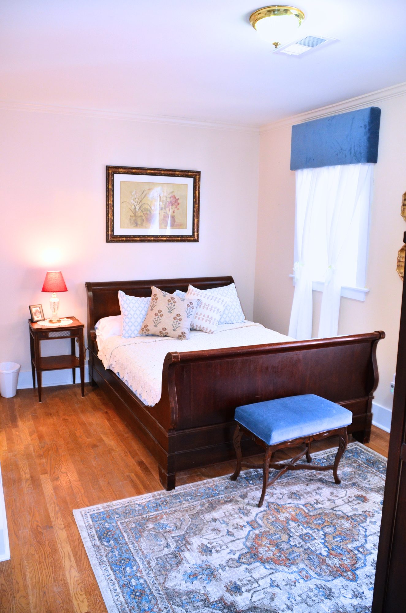 Mary: a dark wood sleigh bed with white linens, a blue upholstered bench at its foot, a blue window valance, and a patterned blue rug.